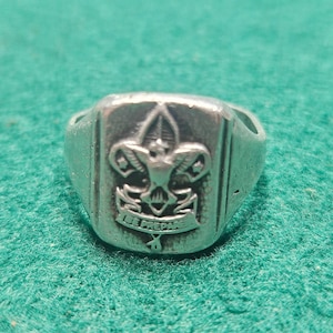 May include: Silver-toned ring with a rectangular face featuring a Boy Scout emblem. The emblem includes a fleur-de-lis, a banner with the words "BE PREPARED," and other details. The ring has a vintage appearance.