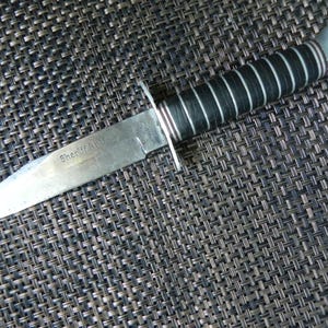 May include: A vintage knife with a silver blade and a black handle. The blade is inscribed with the words "Sheriff Arms". The handle is made of black and white stacked leather. The knife has a silver bolster and a silver end cap.
