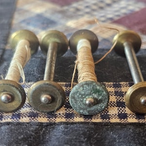 May include: Four antique metal spools with thread. The spools have a brass-colored finish with a central metal rod and circular ends. Two spools are wrapped with off-white thread, and two with tan thread. The spools are on a patterned fabric.