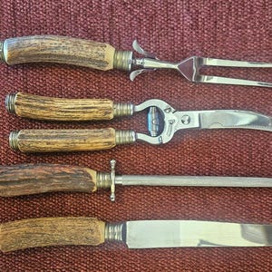 May include: A set of four kitchen tools featuring textured brown antler-style handles with silver-toned metal ferrules. The set includes a two-pronged carving fork, a curved blade tool, a sharpening steel rod, and a straight-edged carving knife.
