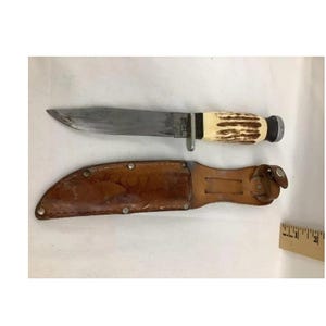 May include: A vintage hunting knife with a worn, silver blade and a handle made of cream and brown material. The knife rests above a brown leather sheath with metal rivets and a belt loop.