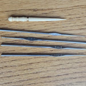 May include: A collection of crochet hooks and a wooden tool are arranged on a wooden surface. The hooks are made of metal and vary in size. The wooden tool has a decorative handle.