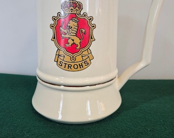 May include: Cream-colored ceramic beer stein with a gold rim and handle. The stein features a red and gold crest with a lion and crown, and the word "STROH'S" in gold lettering.