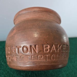 May include: A brown, ceramic jar with a lid. The jar has the words "BOSTON BAKED" and "CHESTERTONS" carved into the side. The jar is sitting on a green surface.
