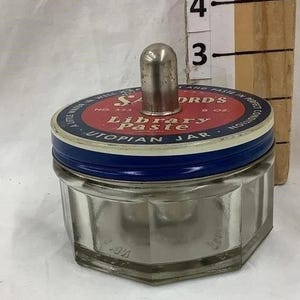 May include: A glass jar with a blue and white lid. The lid has a silver top and the text "S.T. DRDS Library Paste 8 oz. UTOPIAN JAR" printed on it.