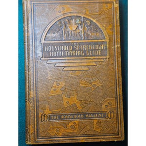 May include: A vintage book titled "The Household Searchlight Homemaking Guide" with a brown cover. The cover features embossed geometric patterns and a detailed illustration of a house and trees. The text "The Household Magazine" is also visible.