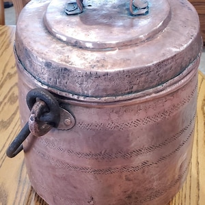 May include: A vintage copper pot with a lid and handle. The pot has a cylindrical shape with etched decorative patterns around the body. The lid has a handle for easy lifting. The pot is sitting on a wooden surface.
