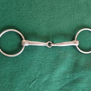 May include: A silver-colored horse bit with two circular rings connected by a central joint and two straight bars. The bit is designed for use in equestrian activities and is set against a green background.
