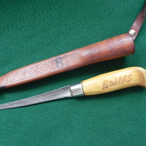 May include: A knife with a wooden handle and a leather sheath. The knife blade is silver and the handle is light brown with the word "BANES" etched into it. The sheath is brown and has a geometric pattern.