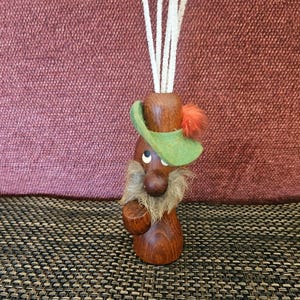May include: A wooden figurine of a gnome with a green hat, a fluffy beard, and a small orange pom-pom. The gnome holds a small wooden cup and has white pipe cleaners in its head.