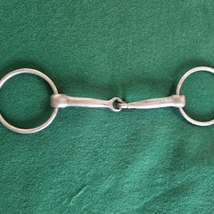May include: A silver-colored metal horse bit featuring two circular rings connected to a central jointed mouthpiece. The bit has a smooth, metallic finish and a classic design with a hinged center section that allows for movement between the two sides.