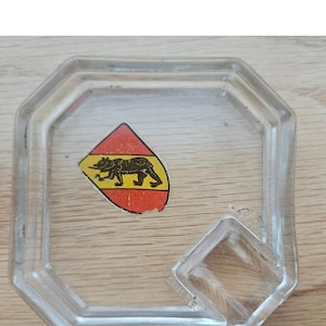 May include: Clear glass ashtray with an octagonal shape and a small, rectangular rest. A colorful sticker with a bear emblem is affixed to the ashtray's surface. The ashtray is set on a wooden surface.