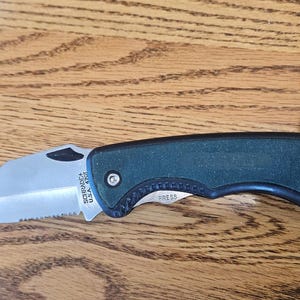 May include: A folding knife with a partially serrated blade and a dark green handle. The blade is silver and has the text "SCHRADE USA 40T". A brown leather strap is attached to the end of the handle.