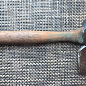 May include: A hand tool featuring a weathered wooden handle with visible grain and a dark, textured metal head. The metal head is securely attached to the handle, showing signs of age and use with a flat, wide blade edge and a blunt poll.