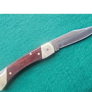 Könnte beinhalten: Ein Klappmesser mit polierter Stahlklinge und einem Griff aus dunklem Holz und Messingakzenten. Das Messer ist geöffnet und zeigt die scharfe Klinge vor einem grünen Hintergrund.