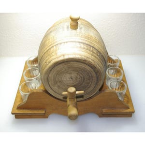 May include: A wooden barrel-shaped liquor dispenser with a spigot, set on a wooden base. The dispenser is surrounded by small shot glasses. The barrel has a light brown finish with a wooden stopper on top. A charming addition to any home bar.