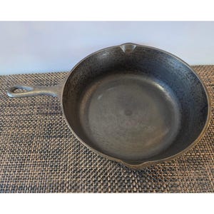 May include: A cast iron skillet with a long handle and a loop at the end. The skillet is dark gray and has a well-worn appearance. The pan is sitting on a textured, brown surface.