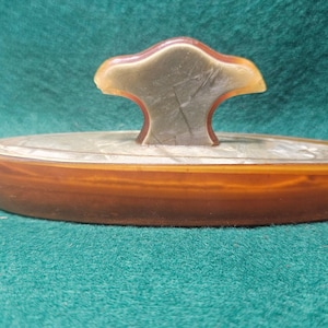 May include: An antique inkwell with a brown, oval base and a decorative handle. The handle is a light brown color with a unique shape. The inkwell is made of a translucent material, and the base has a layered appearance.