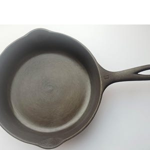 May include: A dark grey cast iron frying pan with a long handle and a rounded cooking surface. The pan has a number 6 stamped on the handle. The handle has a hole for hanging. The pan is designed for cooking.