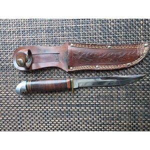 May include: A fixed-blade knife with a polished steel blade and a stacked leather handle featuring red and black spacers. The knife is accompanied by a brown leather sheath embossed with a floral pattern and secured with a metal snap button.