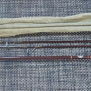 Puede incluir: Caña de pescar con mosca desmontada, con mango de corcho y detalles metálicos, junto con una funda de lona verde claro. La caña es marrón oscuro y plateada, sobre una superficie gris texturizada.