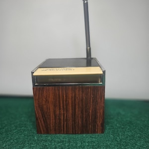 Peut inclure: Une radio portable vintage avec une finition imitation bois sur la partie inférieure et une antenne argentée. La partie supérieure est noire et crème. La radio a une forme rectangulaire et une esthétique rétro.