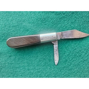 May include: A vintage Barlow pocket knife with a brown handle and two blades. The knife has a silver bolster with the name "BARLOW" engraved on it. The blades are open, showing signs of wear. The knife is on a green surface.