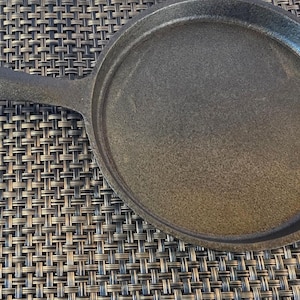 May include: A cast iron skillet with a long handle and a round cooking surface. The skillet is dark gray and appears to be well-seasoned. The handle has a hole for hanging. The skillet is designed for stovetop or oven use.