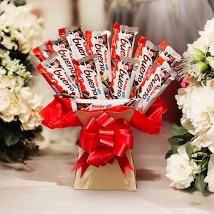 May include: A gift arrangement of Kinder Bueno chocolate bars presented in a gold box, decorated with a large red bow and tissue. The chocolate bars are individually wrapped in red and white packaging. White flowers are in the background.