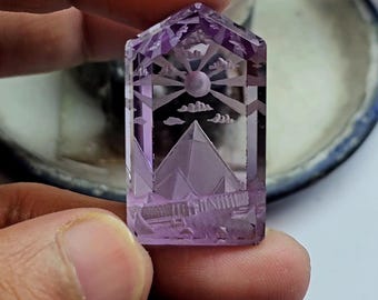 May include: A translucent purple amethyst gemstone carved into a rectangular shape with a pointed top. The interior features an intaglio engraving of a pyramid landscape under a sun with radiating beams and small clouds, set within a frame.