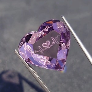May include: A heart-shaped, light purple gemstone featuring a faceted surface with an engraved heartbeat line pattern and a small heart symbol in the center. The stone is held by silver-toned metal tweezers, showcasing its translucent, polished finish.