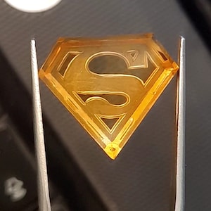 Könnte beinhalten: Ein gelber, diamantförmiger Anhänger mit einem erhabenen, geätzten Superman-Logo.