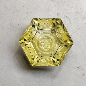 May include: A yellow faceted gemstone with a carved design. The gemstone is shaped like a hexagon and has a intricate pattern on the surface.