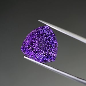 May include: A triangular-shaped purple gemstone featuring an intricate, laser-cut lattice pattern across its surface. The stone displays a deep violet hue with geometric cutouts that create a complex, web-like structure throughout the entire piece.