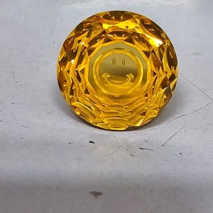 May include: A faceted, round, yellow gemstone with a smiley face etched in the center. The gem has a brilliant cut, reflecting light. The image is a close-up, highlighting the details of the stone.