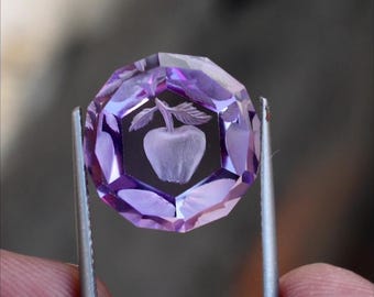 Può includere: Una gemma rotonda, traslucida e di colore viola, caratterizzata da molteplici sfaccettature geometriche lungo il perimetro. Al centro è presente un intaglio inciso che raffigura una mela con due foglie, con una superficie lucida e riflettente.