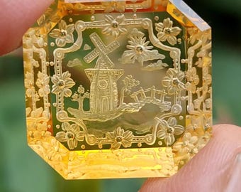May include: An octagonal amber-colored gemstone featuring an intricate intaglio carving of a windmill, a small bridge, and surrounding floral motifs. The transparent yellow stone has beveled edges and a polished surface with detailed engravings.