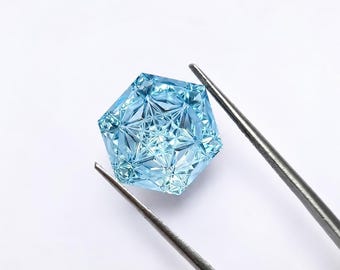 May include: A light blue, hexagonal gemstone featuring a complex, geometric, star-like faceted pattern on its surface. The transparent, icy-colored stone is held by a pair of thin, silver-toned metal tweezers against a plain white background.