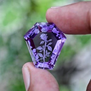 Puede incluir: Una piedra preciosa morada con forma de ataúd que presenta un grabado en hueco detallado de una rosa con hojas y tallo. La piedra violeta translúcida tiene bordes facetados que captan la luz, resaltando el diseño floral en su interior.
