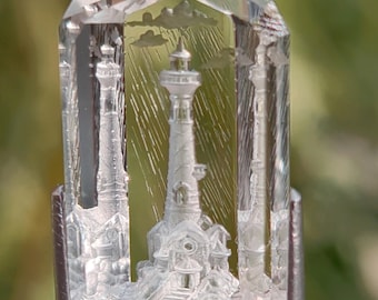 May include: A clear, faceted gemstone featuring an intricate internal carving of a lighthouse scene. The design depicts a central lighthouse tower surrounded by rain, clouds, and rocky terrain, with additional lighthouse silhouettes visible on the sides.