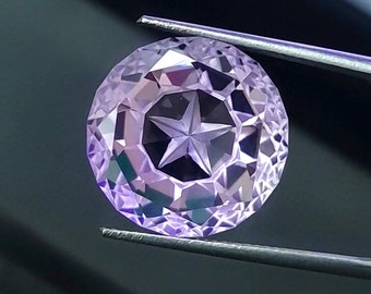 Può includere: Una gemma rotonda color viola chiaro caratterizzata da un motivo a stella tagliato con precisione al centro. La pietra presenta molteplici sfaccettature triangolari e trapezoidali lungo il perimetro, creando una superficie complessa e riflettente.