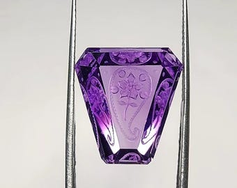 May include: A faceted purple gemstone with a trapezoidal shape, featuring an intricate intaglio carving of a floral paisley motif on its surface. The translucent violet stone is held by silver-toned metal tweezers, showcasing its clear, polished finish.
