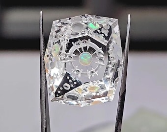 May include: A clear, faceted gemstone featuring an intricate internal design with a central circular opal, radiating spokes, and scattered iridescent opal accents embedded within the transparent material, held by metal tweezers.