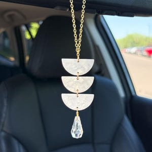 May include: A car rearview mirror hanging ornament featuring a gold-toned chain connected to three stacked white marbled acrylic semi-circle shapes, finished with a clear, faceted, teardrop-shaped crystal pendant dangling at the bottom.