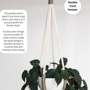 Op de afbeelding: Een witte plantenhanger met een dubbel koordontwerp en grijze accenten, opgehangen aan een bronzen metalen ring. De hanger bevat een witte pot met een donkergroene bladplant. De tekst op de afbeelding luidt: Double Cord Version.