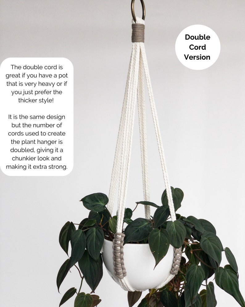 Op de afbeelding: Een witte plantenhanger met een dubbel koordontwerp en grijze accenten, opgehangen aan een bronzen metalen ring. De hanger bevat een witte pot met een donkergroene bladplant. De tekst op de afbeelding luidt: Double Cord Version.