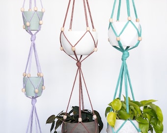 May include: Three macrame plant hangers suspended from metal rings. The left hanger is lavender with three small light blue pots. The center is terracotta with two pots, one white and one dark gray. The right is teal with two pots, one white and one speckled.