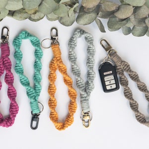 May include: Five macrame wristlet keychains in magenta, teal, mustard yellow, light gray, and taupe. Each keychain features a twisted cord design with a metal lobster clasp and a split ring. One taupe wristlet is attached to a black car key fob.