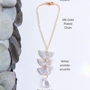May include: A necklace featuring an 18k gold plated chain with a lobster clasp closure. The pendant consists of three stacked white acetate semi-circle accents and a single clear real crystal drop. Text labels identify the components.