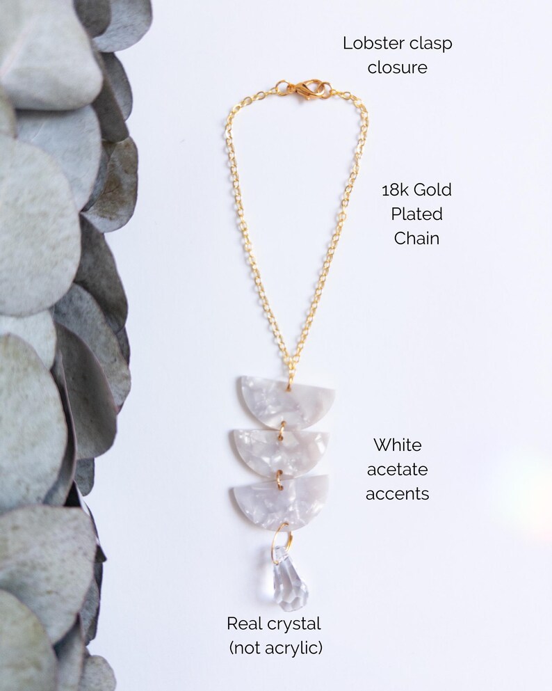 May include: A necklace featuring an 18k gold plated chain with a lobster clasp closure. The pendant consists of three stacked white acetate semi-circle accents and a single clear real crystal drop. Text labels identify the components.